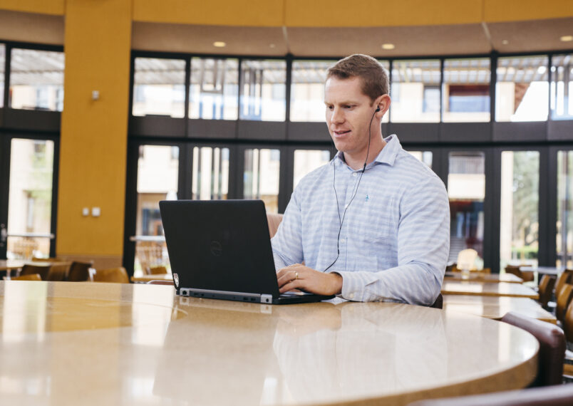 Stanford GSB student participating in virtual class on laptop with headphones in campus common area.