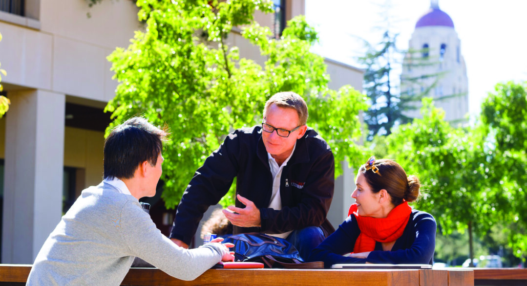 Three individual people conversing while sitting at a picnic table with Hoover Tower in the background.