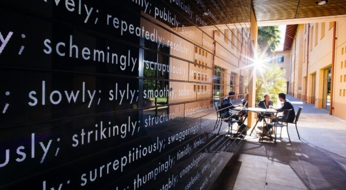 Stanford GSB students collaborating outdoors with artistic word wall installation and sunlight in background.